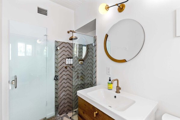A modern bathroom with a glass shower enclosure, a white sink with a gold faucet, and a round mirror on the wall.