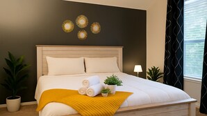 Un lit bien fait, avec du linge de lit blanc, un plaid de couleur moutarde et des serviettes enroulées. Deux plantes en pot sont placées de chaque côté du lit.
