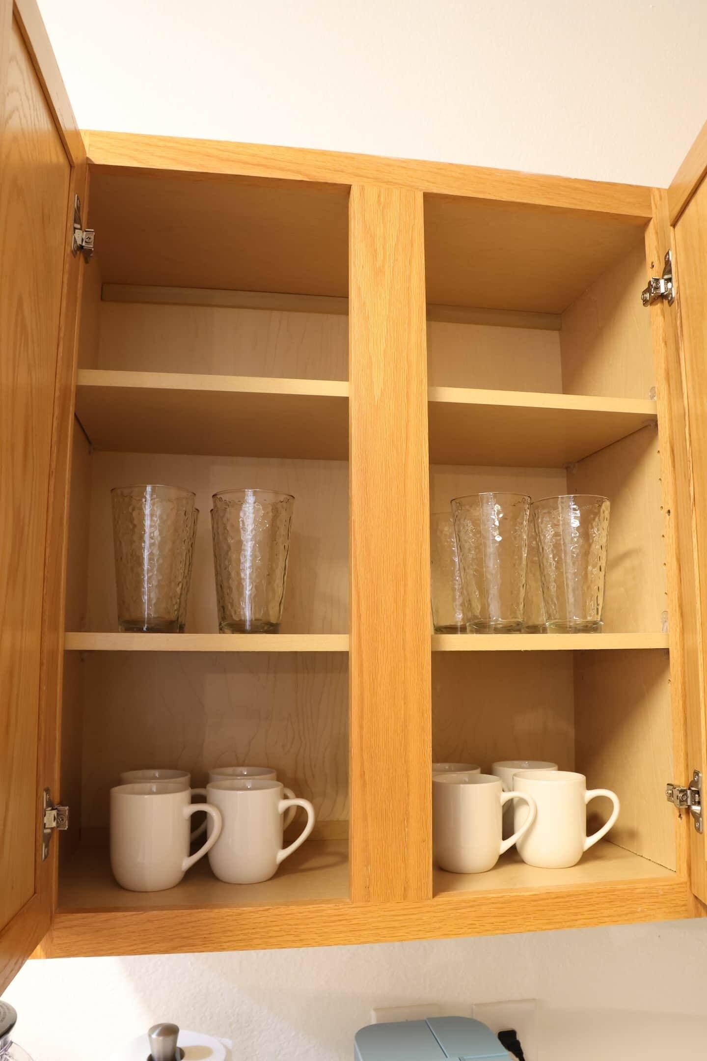 A wooden cabinet with two shelves; the top shelf holds two clear glass cups, and the bottom shelf holds four white mugs.