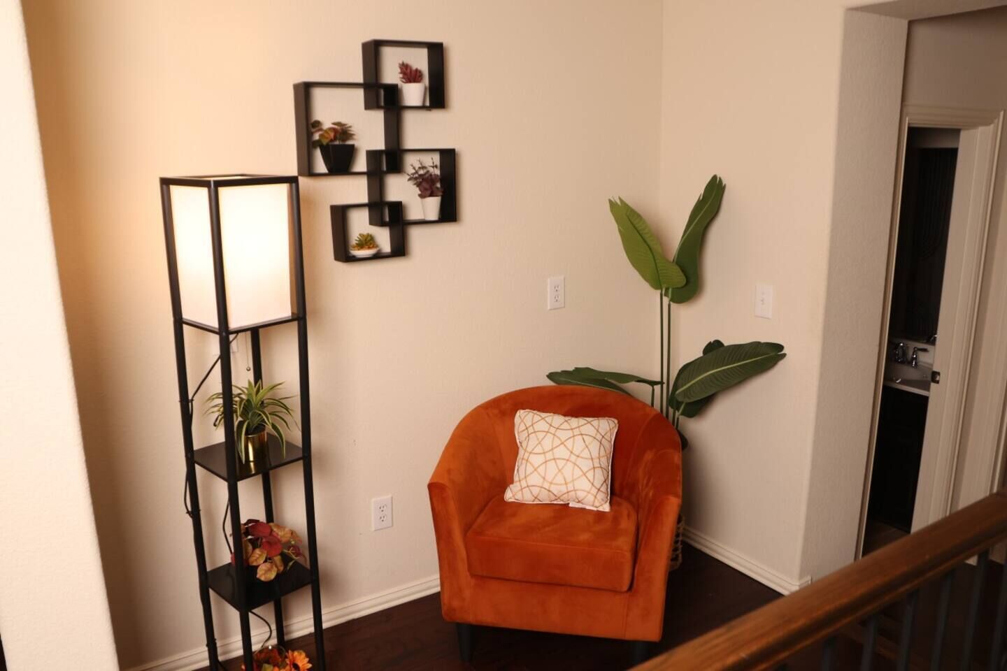 A corner of a room with a tall black shelving unit, an orange armchair with a patterned cushion, and a large green plant.