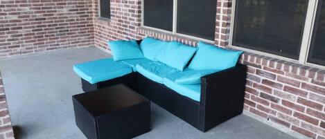 A black wicker sectional sofa with turquoise cushions and a matching black coffee table, positioned on a concrete patio against a brick wall with large windows.