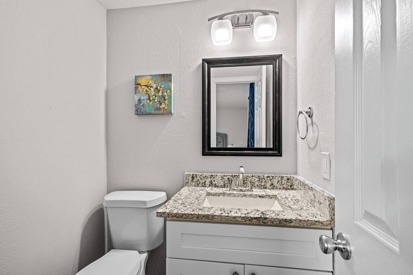 Un baño con lavamanos de encimera de granito, inodoro blanco, espejo pequeño enmarco y lámpara de pared.