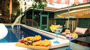 Een zwembadterras met een houten dienblad vol fruit, een cocktail en een ligstoel.
