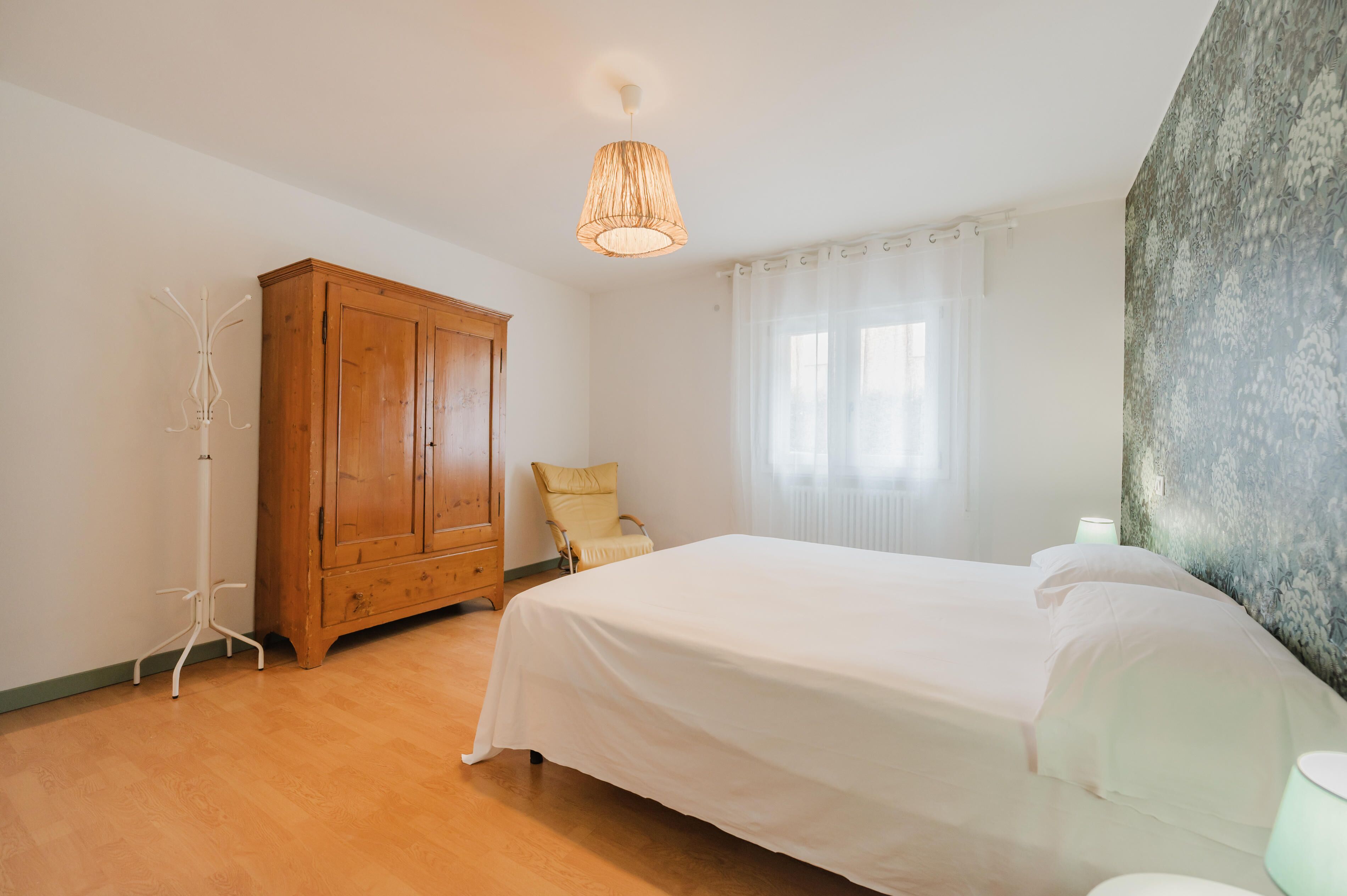 A bedroom with a wooden wardrobe, a bed with white bedding, a chair, and a hanging light fixture.