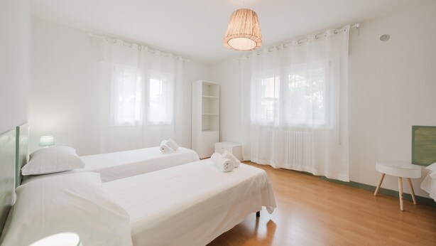 A hotel room with two beds, a small table, a shelf, and a hanging light fixture.