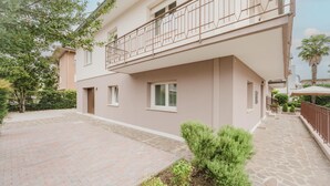 A two-story residential building with a balcony, paved courtyard, and surrounding greenery.