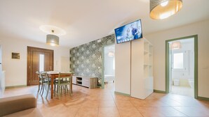 A modern living room with a dining area, a TV mounted on the wall, and a kitchen visible through an open door.
