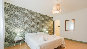 A bedroom with a floral wallpaper, a bed with white linens, two bedside tables with lamps, and a hanging light fixture.