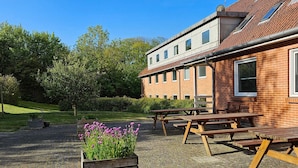 A building with a brick exterior, multiple windows, and a tiled roof. There are outdoor wooden picnic tables and a planter with purple flowers. - 2 Person Holiday Park Home in Lemvig (Lemvig)