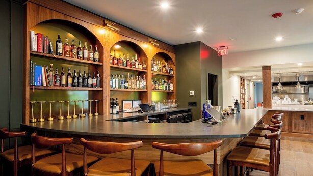 A bar area with wooden shelves stocked with liquor bottles, a bar counter, and leather bar stools. - The Nightingale (Lewiston)