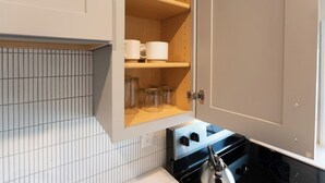A kitchen with a white tiled backsplash, a stove with a kettle, and a wooden cabinet with shelves holding glass jars and a roll of paper towels. - The Nightingale (Lewiston)