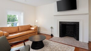 A living room with a leather sofa, a round coffee table, a fireplace, and a flat-screen TV mounted above it. - The Nightingale (Lewiston)