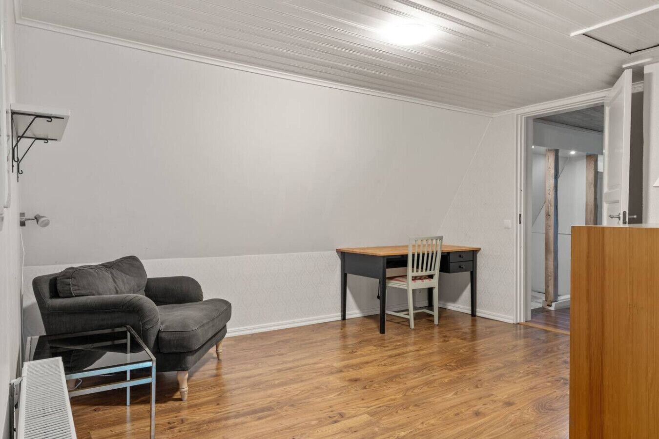 Une pièce avec un plancher en bois, un canapé noir, une table à plateau en verre, un bureau en bois avec une chaise, et un mur blanc.