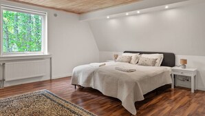 A bedroom with a sloped ceiling, a large window, a bed with a headboard, bedside tables, and a radiator.