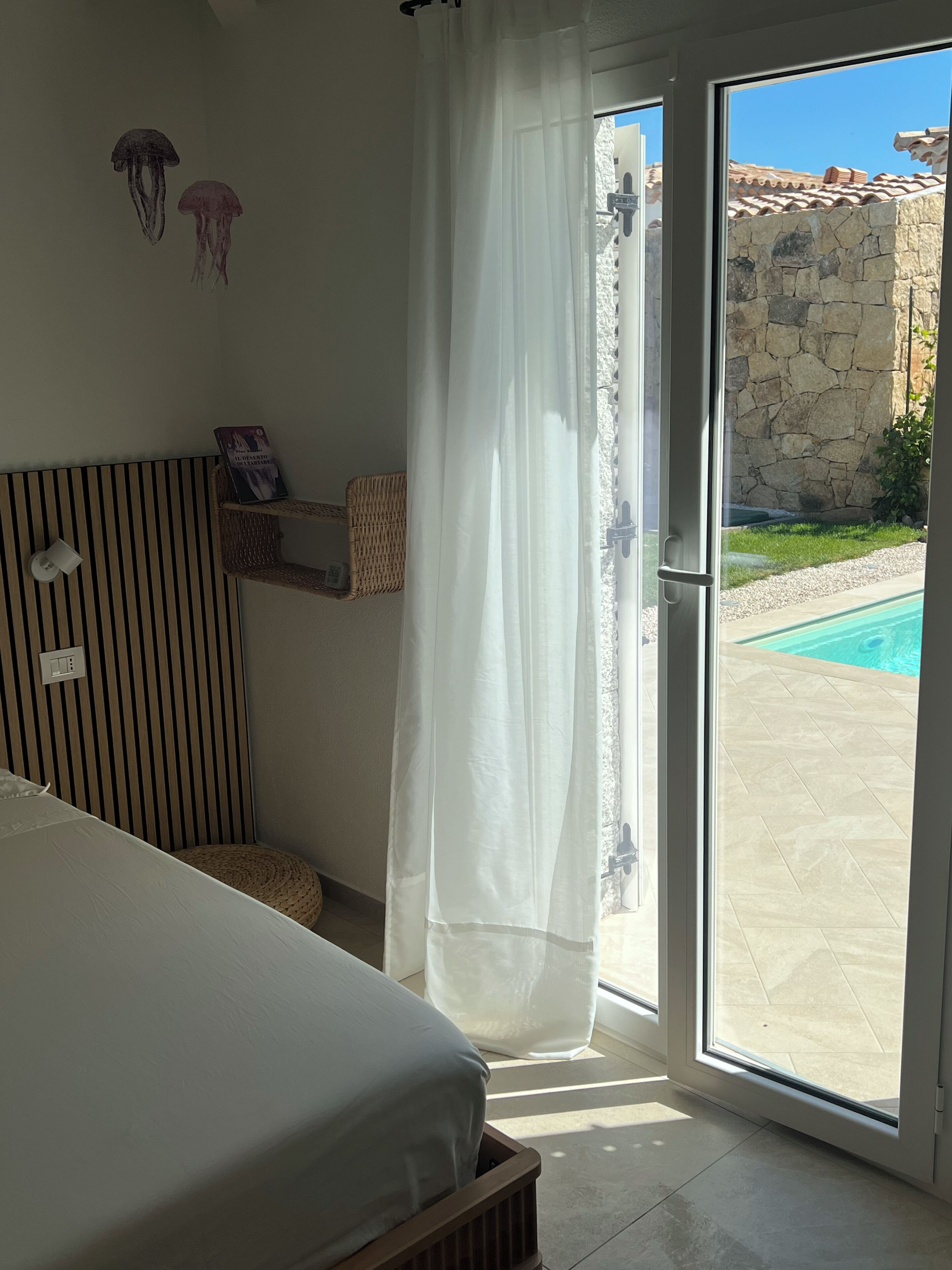 A bedroom with a bed, a window with a curtain, a wall-mounted shelf with a book, and a view of an outdoor swimming pool.