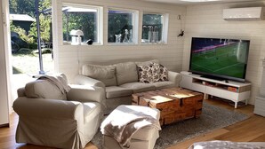 Ein Wohnzimmer mit beiger Couch, Couchtisch und Fernseher. Fenster mit Vorhängen, ein Kamin und Blick ins Grüne sind ebenfalls zu sehen.