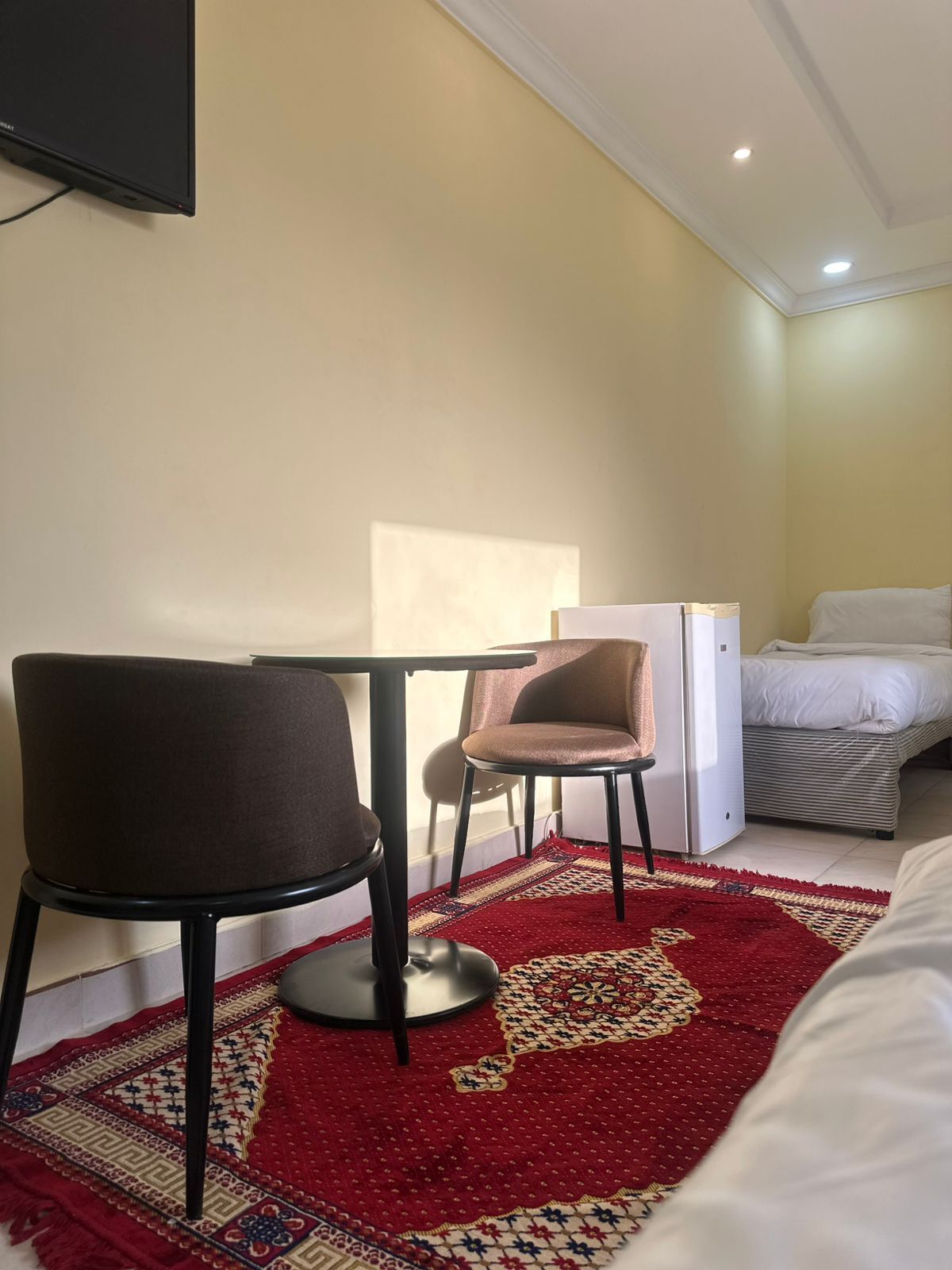 Et hotelværelse med en seng, et lille bord med to stole, et køleskab og et tv ophængt på væggen.