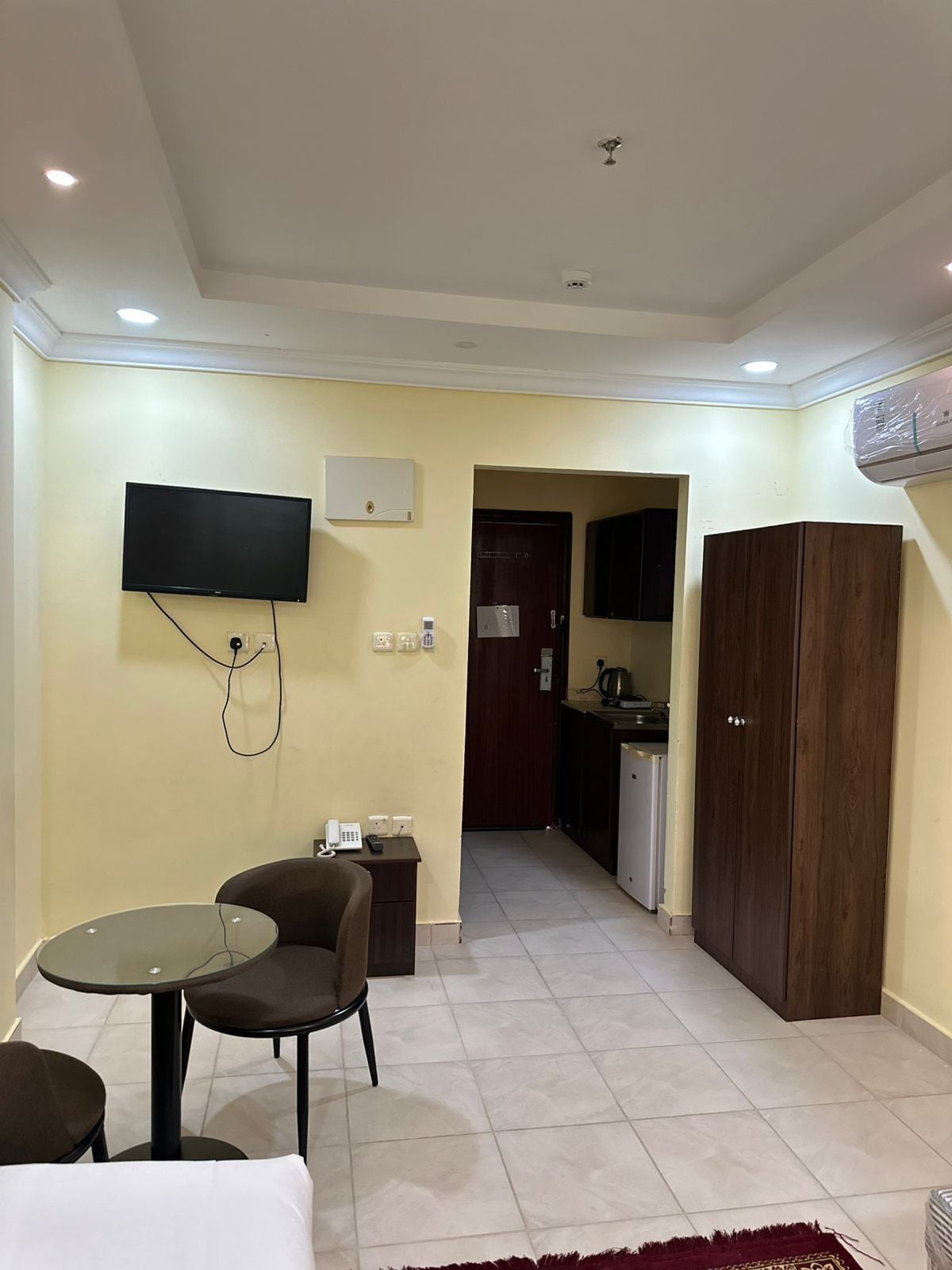 Habitación de hotel con televisor montado en la pared, una pequeña mesa con sillas, un armario y una cocina visible a través de una puerta abierta.