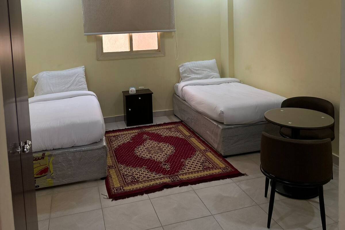 A hotel room with two beds, a small table, a window with a blind, a red patterned rug, and a ceiling light.