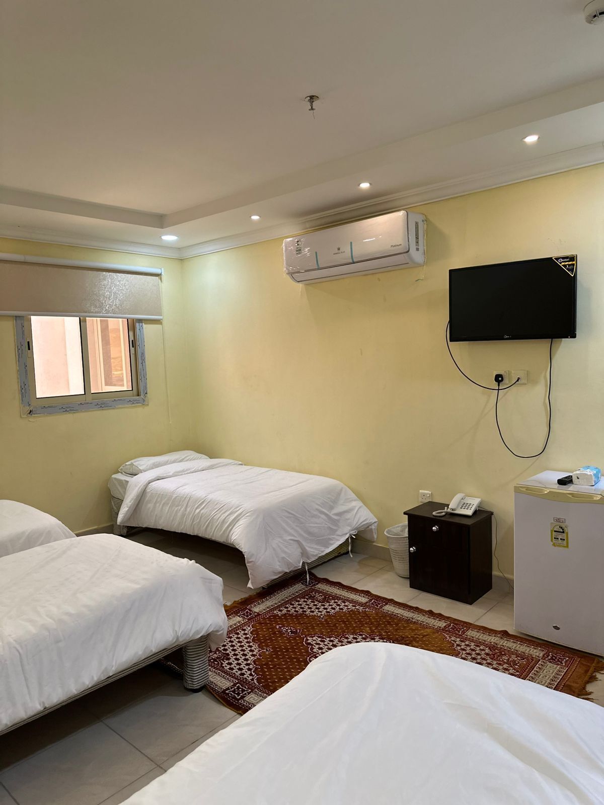 A hotel room with two single beds, a TV mounted on the wall, an air conditioning unit, a small desk with a phone, and a patterned rug.