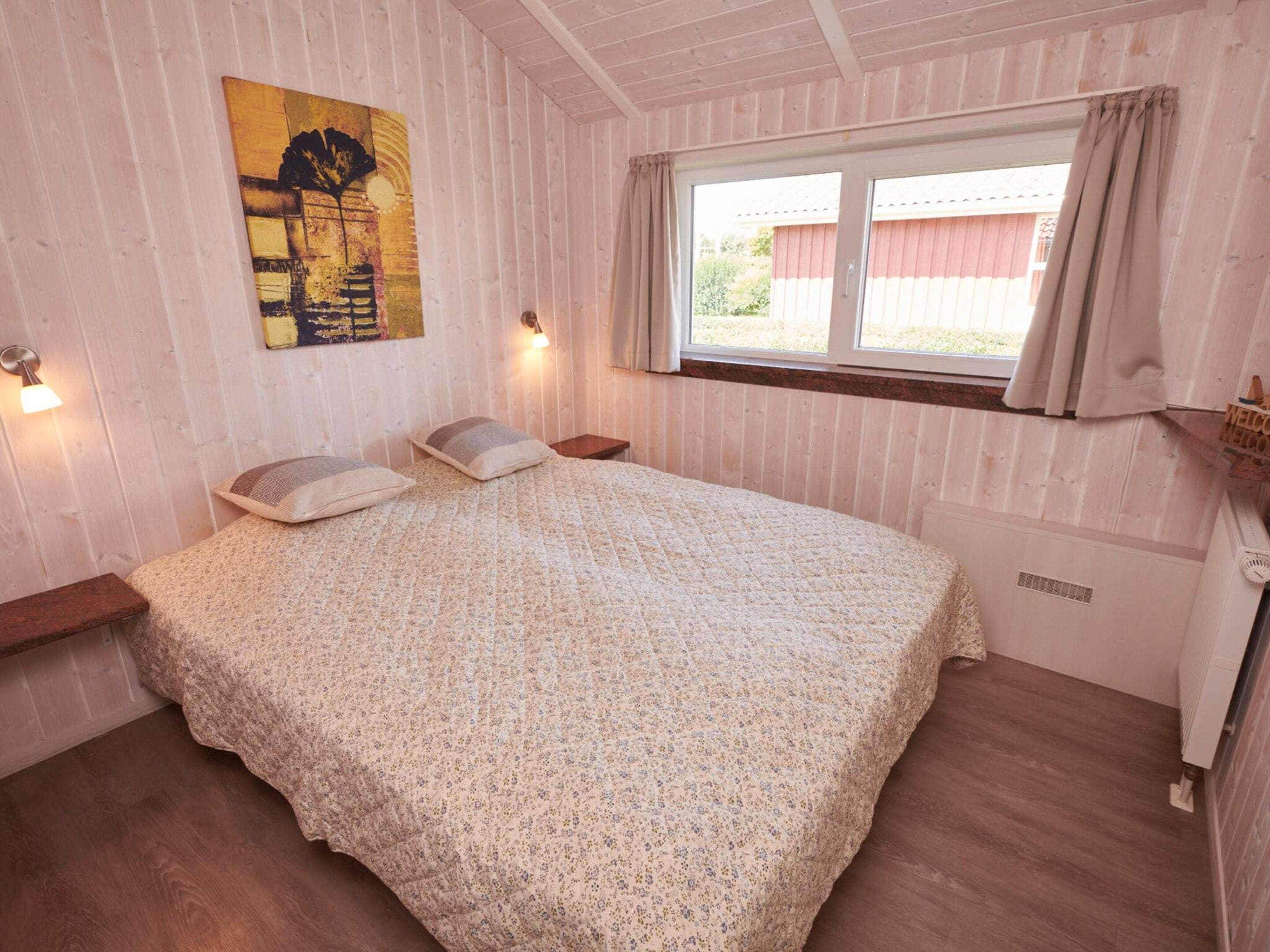 Een slaapkamer met een bed, twee kussens, een nachtkastje, een wandlamp, een raam met gordijnen en een radiator.