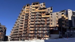 Un edificio de varios pisos con balcones, una pista de esquí en primer plano y un cielo azul despejado.