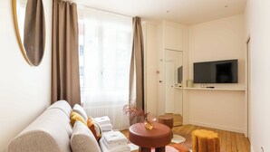 A modern living room with a sofa, a TV, a small round table, and a window with curtains. - Port-royal Residence - Modern Apartement-1br- 4P (Paris)