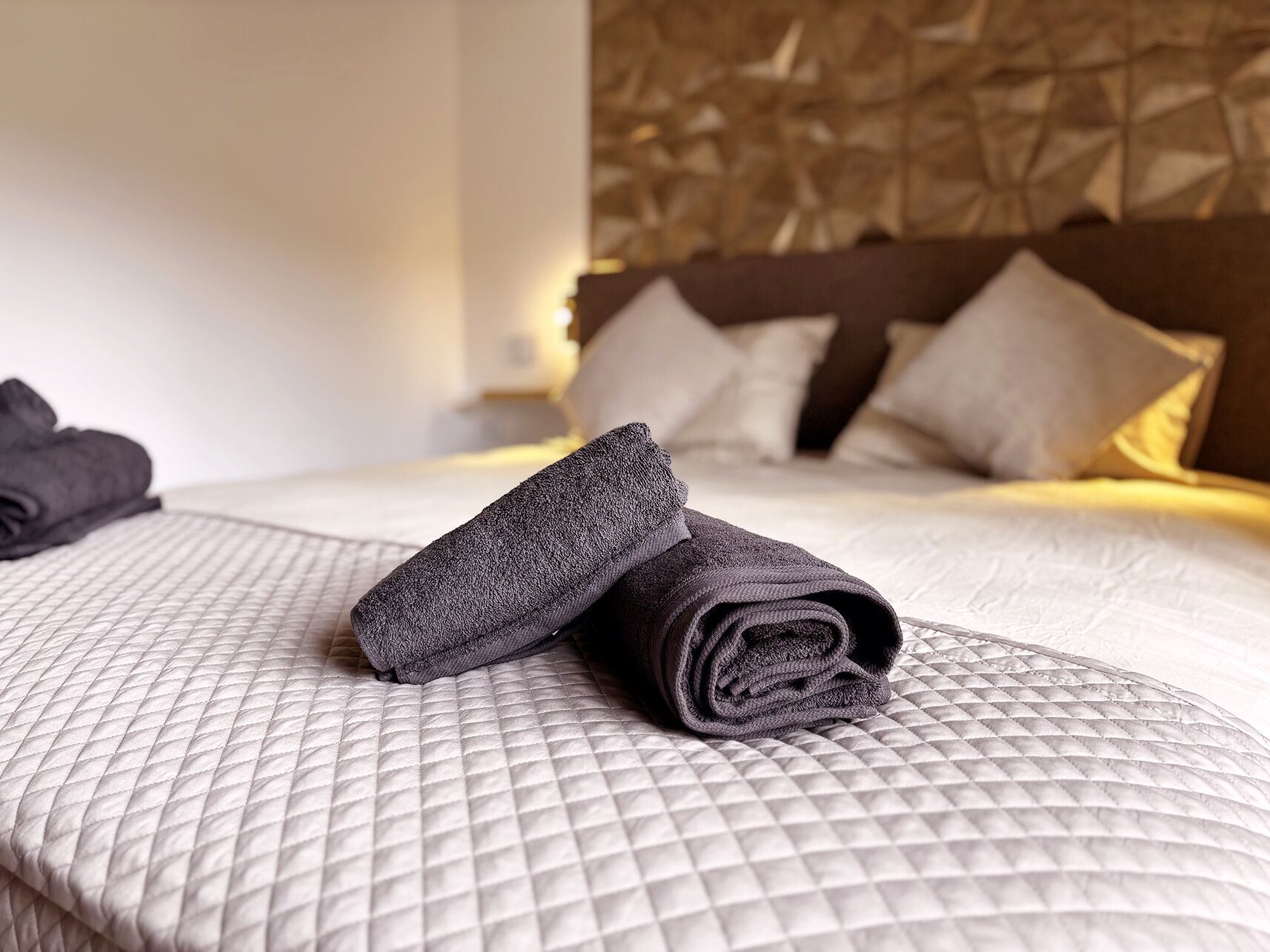 A neatly made bed with a quilted cover, pillows, and a folded towel on the bed.