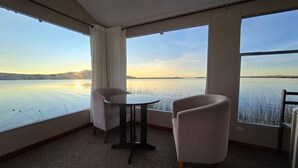 A room with two chairs and a table, large windows with a view of a calm lake and distant mountains. - Uros Maya lodge (Puno)