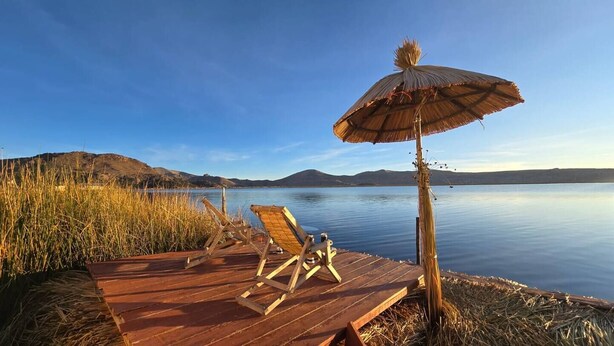 A wooden deck with two lounge chairs and a thatched umbrella by a calm lake with distant hills. - Uros Maya lodge (Puno)