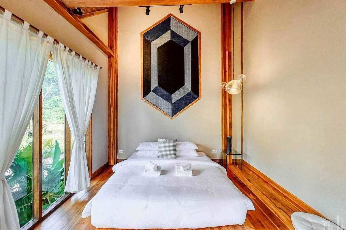 Une chambre avec un grand lit, un plancher en bois, une décoration murale aux motifs géométriques et une vue sur la verdure par la fenêtre.