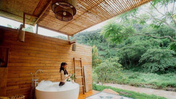 A wooden cabin with a large bathtub filled with bubbles, a ladder, and a view of a lush forest.