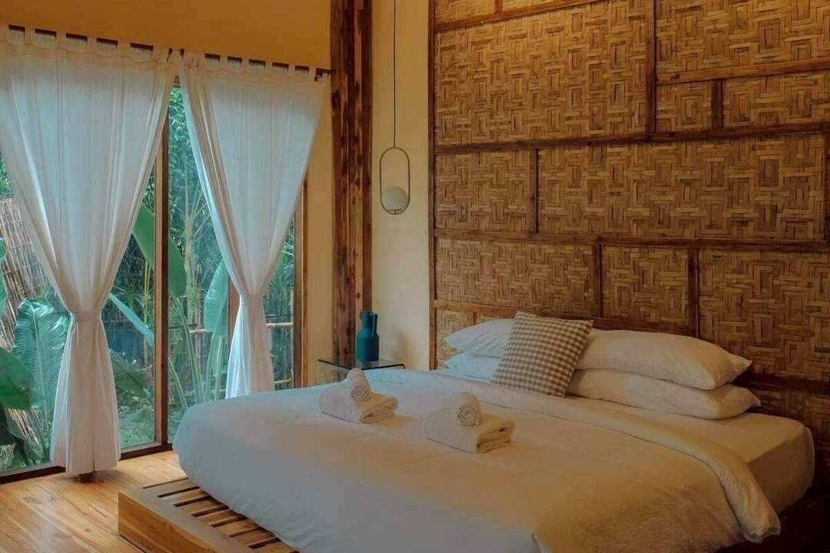 Une chambre à coucher avec un lit, des rideaux, un mur orné d’un motif tissé et une vue sur la verdure à l’extérieur.