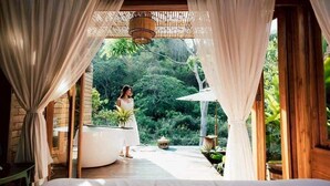 A bedroom with a bed, a large window with curtains, a wooden ceiling, and a view of greenery outside.