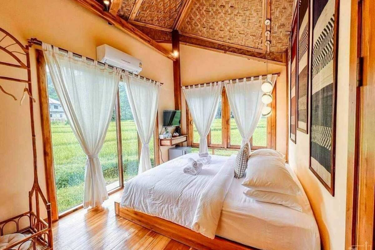 Une chambre avec un lit en bois, du linge de lit blanc, une télévision et de grandes fenêtres avec des rideaux.