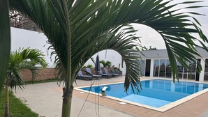 A swimming pool with a tiled deck, surrounded by lounge chairs and palm trees. - Alojamento Santos Silva  (São Tomé)