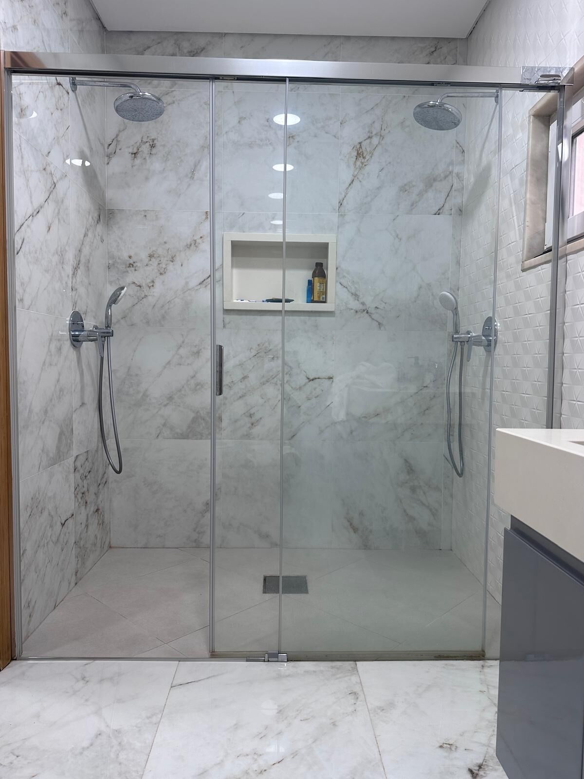 A modern bathroom with a glass-enclosed shower, marble tiles, and a small shelf with toiletries.