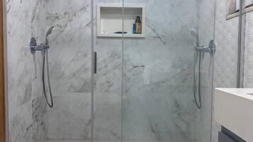 A modern bathroom with a glass-enclosed shower, marble tiles, and a small shelf with toiletries.