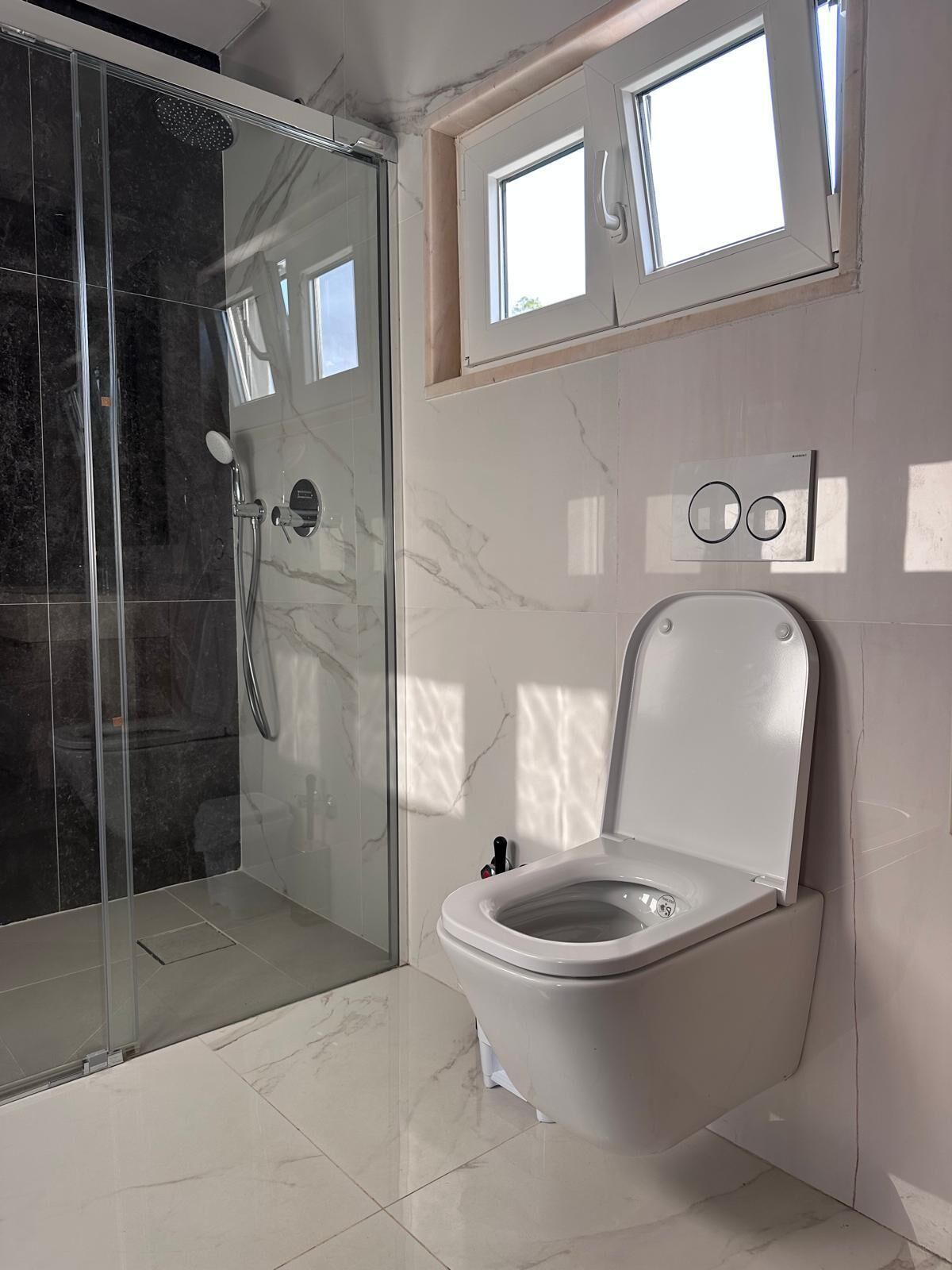 A modern bathroom with a glass-enclosed shower, a toilet with a bidet attachment, and a window above the toilet.