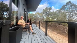 En træterrasse med to personer siddende, hvorfra der er udsigt over et landskab med træer og en klar himmel.