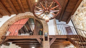 A rustic interior with exposed wooden beams, a large wooden wheel chandelier, a stone wall, and a balcony with a metal railing.