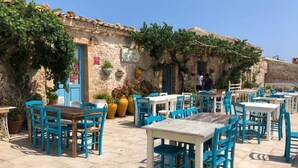 An outdoor café with tables and chairs, a stone building with a blue door, and potted plants.