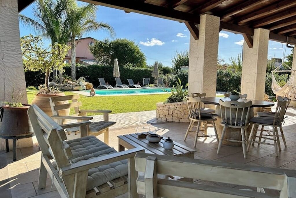 Een overdekt terras met houten meubels, een zwembad en een uitzicht op een huis op de achtergrond.