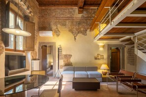 Un salón amplio con un sofá grande, una mesa de centro de vidrio y una televisión. Se ve una escalera con barandilla de madera que sube a un nivel superior.