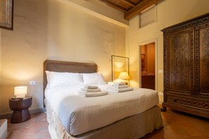 Una cama bien hecha con sábanas y toallas blancas, una lámpara de noche y un aparador de madera con intrincados grabados.