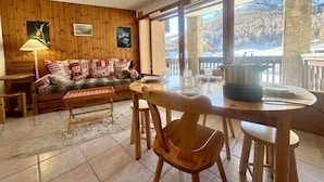 A cozy living room with a wooden sofa, a dining table with chairs, and a view of snow-covered mountains through large windows.