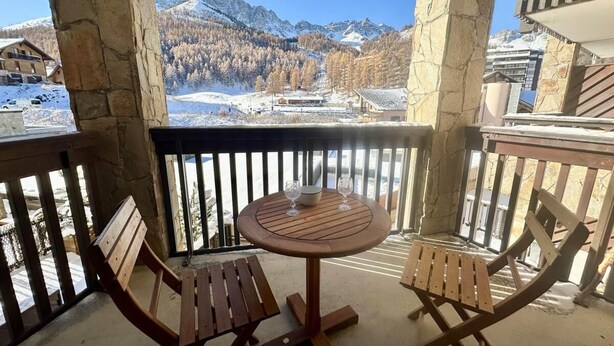 A balcony with a wooden table and two chairs, overlooking a snowy mountain landscape.