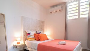 A neatly made bed with a red blanket and two colorful pillows, a bedside lamp, and a wall-mounted air conditioner. - Villa Angerville, spacious villa that can accommodate up to 24 people (Le Moule)
