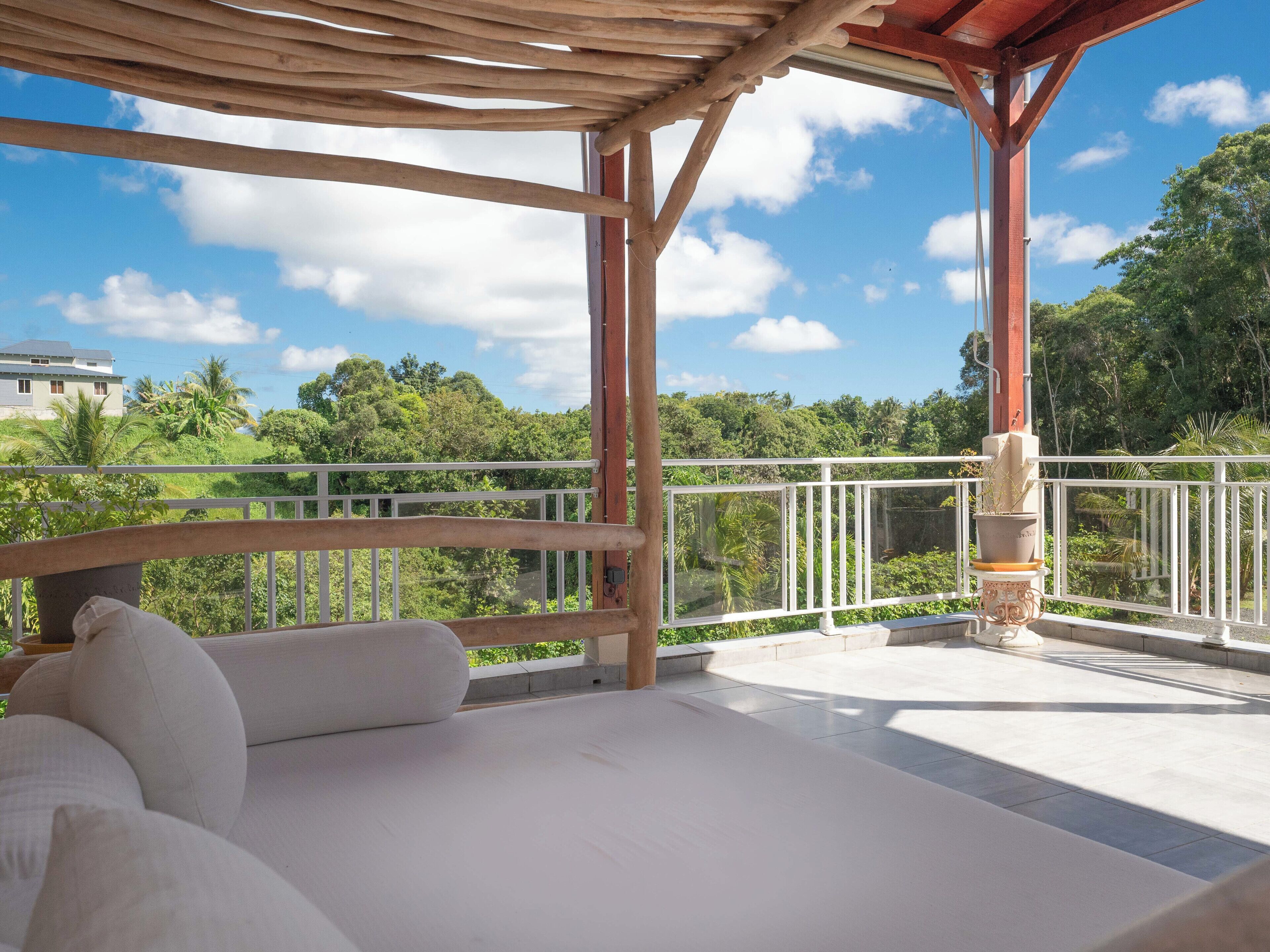 A balcony with a wooden pergola, white cushioned seating, and a view of lush greenery and a distant building.