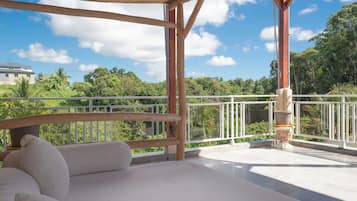 A balcony with a wooden pergola, white cushioned seating, and a view of lush greenery and a distant building.